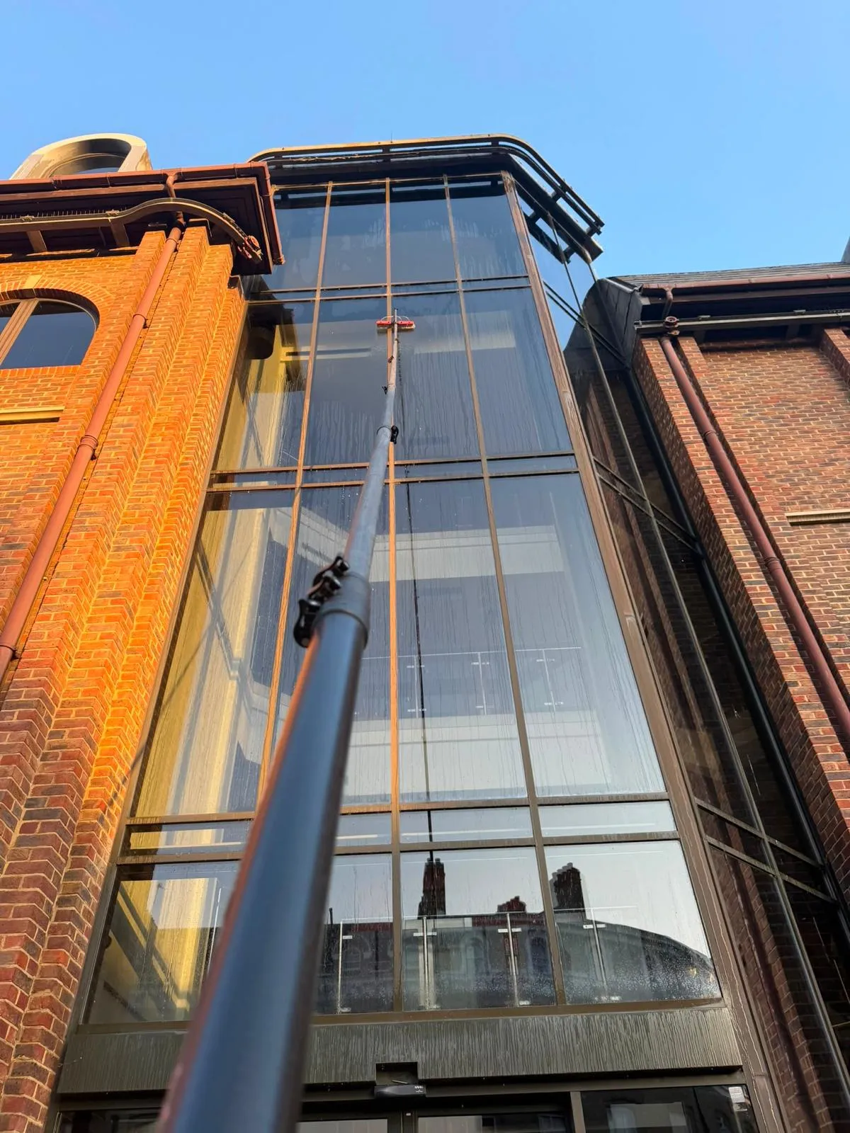 Looking up at a full-height glazed atrium between two red-brick commercial buildings, being cleaned with an extended water-fed pole