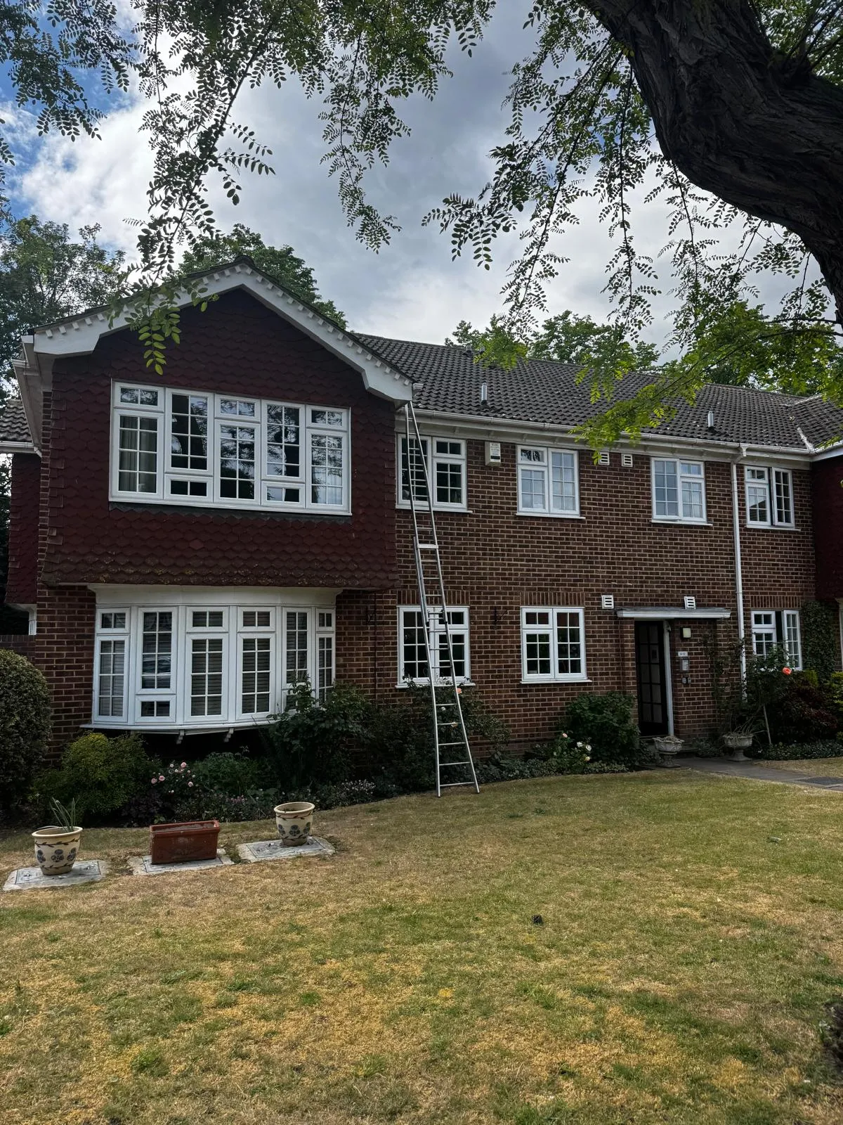Large tile-hung red-brick family home with white bay windows, a ladder set up for the upper windows and a lawn to the front